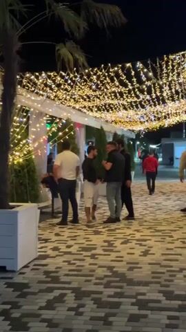 The video shows an outdoor area at night, decorated with numerous string lights suspended from an overhead canopy. Several palm trees are visible, one prominently in the foreground in a white planter. People are gathered in small groups, some appearing to be conversing. A bar area is visible in the background with people standing and sitting. A scooter is parked in the lower left portion of the frame. The ground is paved with a pattern of dark and light colored rectangular stones. The atmosphere appears to be social and lively, with the lighting suggesting an evening entertainment venue.