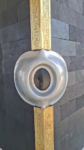 The video shows a close-up view of the Kaaba in Mecca, Saudi Arabia. The camera pans across the black stone facade, highlighting the gold-plated decorations and the Hajr-e-Aswad, or Black Stone, embedded in one of the corners. The sun shines brightly in the sky, casting shadows and illuminating the building. In the background, a large white canopy provides shade, and a golden lantern stands on a pedestal. The surrounding area appears to be an open courtyard with some buildings and vegetation visible. The overall scene is one of religious significance and architectural beauty.