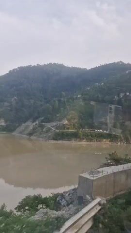 A murky river flows through a valley flanked by tree-covered hills. The sky is overcast. The camera is positioned on a roadside barrier, possibly indicating the footage was captured from a moving vehicle.