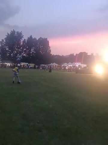 A wide outdoor shot at sunset shows a large grassy field with a fair or festival in the background. Several tents and food trucks are visible, along with numerous people strolling around. A child in a yellow shirt and blue pants runs across the field. The sky is a mix of purple, pink, and orange from the sunset, with a bright light source on the right. Trees line the background, and distant mountains are faintly visible. Lively music plays.