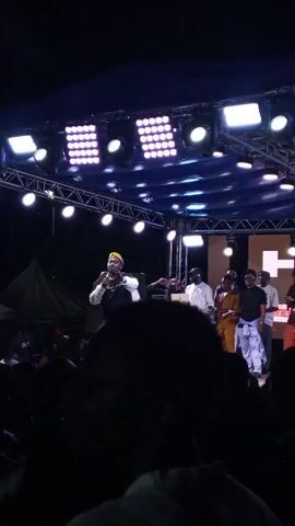 A video shows a man in a white and black outfit and a gold head wrap holding a microphone on a stage. He is speaking and gesturing with his free hand. There are several other people on the stage behind him, some appearing to be musicians with instruments. A large crowd is visible in the foreground, silhouetted against the bright stage lights. The stage is decorated with lights and a large screen in the background displays the letters "HO". The event appears to be an outdoor concert or performance at night. The man on stage is engaging with the audience.