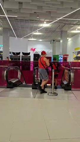 The video shows the interior of a shopping mall. In the foreground, there are escalators with pink railings. People are seen using the escalators. In the background, there is a cinema with a sign that reads "КИНОТЕАТР". Further in the background, there are food stalls, including a Burger King. People are sitting at tables, eating and talking. The overall environment appears to be a busy, indoor public space.