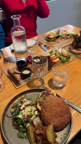 The video shows an indoor dining scene, likely in a restaurant, featuring a table set with multiple dishes and drinks for several people.

In the foreground, a large, dark grey plate holds a brown, bun-shaped item, roasted potato wedges, and a fresh green salad with various vegetables such as cucumber, radish, red onion, and crumbled white cheese or feta.

Further back on the wooden table, there are several clear glass platters. One on the middle right holds a salad with cherry tomatoes and a burger patty or cutlet. Another on the far right displays a similar salad accompanying a substantial piece of meat, possibly a lamb shank, with a bone protruding and a rich sauce. To the left, a rectangular glass platter features several small, golden-brown pastry-like items or breadsticks, accompanied by three small ramekins containing dark and light dips or sauces.

Drinks include a glass carafe of water with condensation on the left, and several clear drinking glasses, some filled with water and ice, along with a stemmed glass. Cutlery (forks and knives) is visible on the table.

Two individuals are partially visible: one wearing a red top is seated on the far side of the table, and another person's arm and part of a blue top are seen on the left. The lighting is warm and inviting. The scene suggests a meal is being enjoyed or is about to be. No text is visible in the video.