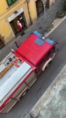 A red fire truck is parked on a cobblestone street in front of a building with a yellow facade. A person wearing a white shirt and a red apron is visible inside the building, which has a sign that reads "MACELLERIA". Next to the fire truck, three individuals in orange and white uniforms stand outside a doorway. Further down the street, another red emergency vehicle is parked, with several firefighters in uniform gathered around it. Two ambulances and a black car are also present on the street. The scene appears to be a street emergency response.
