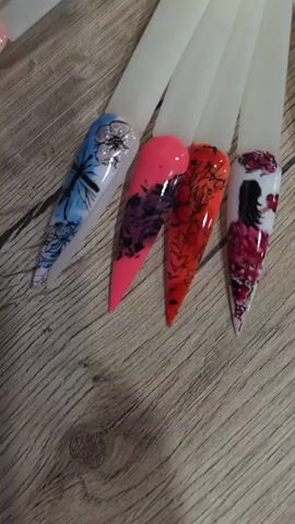 The video displays a collection of false nails, arranged on a wooden surface. Each nail is mounted on a white plastic holder and features intricate designs. Some nails are adorned with floral patterns, while others showcase abstract designs, lips, and a tropical sunset scene. The lighting is bright, highlighting the details of the nail art. The overall impression is that of a display of nail art samples.