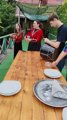 A group of people are gathered around a wooden table outdoors. Two young women in traditional red and black outfits are playing musical instruments. One woman plays a large drum, while the other holds a small, brown instrument. A young man stands next to the woman playing the drum, looking towards the camera. A woman in a colorful striped shirt reaches towards the table, holding a glass. The table is set with white plates, silverware, napkins, and several wine glasses. The background shows greenery and a building with pink and white accents. The scene appears to be a celebration or gathering.