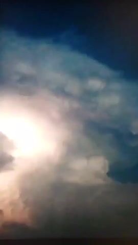 This is a vertical video of a stormy sky with lightning. Dark, ominous clouds fill the frame, with streaks of white and grey. A bright flash of lightning illuminates the center of the image, casting a pinkish glow on the surrounding clouds. The bottom of the frame shows a dark, indistinct horizon line.