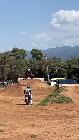 The video captures a motocross training session set against a backdrop of a partly cloudy sky and distant mountains. Two riders on dirt bikes navigate the terrain. One rider wearing white and black gear remains on the ground level. The other, clad in a yellow helmet and darker gear, performs jumps over the dirt mounds. Several vans are parked in the distance, between the terrain and the treeline. The dirt bike track is located beneath powerlines strung between utility poles. The ground cover is primarily dirt, with small patches of grass.