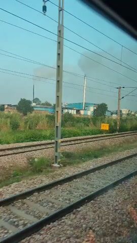 The video shows a view from inside a moving train. Train tracks are visible in the foreground and background. On the left side of the tracks, there is overgrown vegetation and some buildings with industrial smoke plumes rising into the sky. A pole with overhead electrical wires for the train is also visible. A blue sign with yellow lettering, reading "1402/4", is affixed to a pole in the foreground. The sky is clear with some clouds. The overall impression is a rural or semi-industrial landscape seen from a moving train.