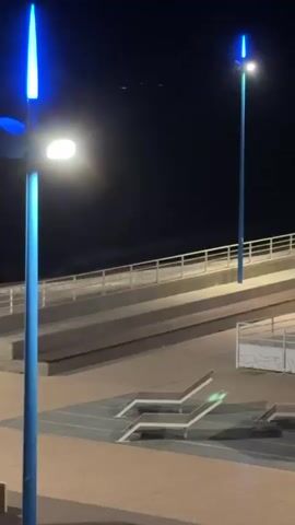 The video shows a night scene with artificial lighting. Two tall, blue streetlights are visible, one in the foreground and one in the background. The foreground streetlight has a bright white light illuminating downwards, while the upper part of the light has a blue glow. The background streetlight is slimmer and also emits a bright white light, with a similar blue accent at the top. In the middle ground, there's a raised walkway with a metal railing. Below the walkway, there are tiered concrete structures. In the foreground, on a paved surface, there are several lounge chairs or benches, some of which have green lights on them. The sky is dark, indicating nighttime. The overall impression is of an urban or recreational area illuminated for nighttime use.