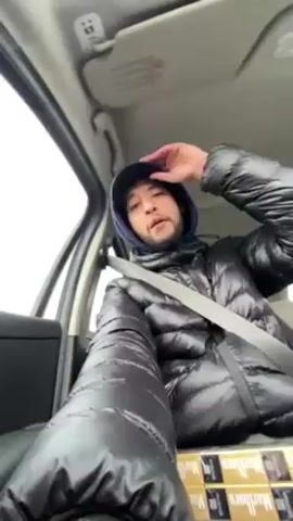 The video shows a man in a car wearing a puffy black jacket, a dark baseball cap, and a light blue hoodie underneath the jacket. He is wearing a seatbelt and appears to be rapping or mouthing words to music playing in the car. In the lower part of the frame there are numerous packs of Marlboro cigarettes visible. The video is shot from a low angle from the passenger seat. It looks like it could be overcast outside.