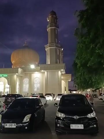 The video shows a mosque with a prominent dome and minaret at dusk. The mosque is illuminated by artificial lights, and the sky is a deep blue with some clouds. In the foreground, there is a parking lot filled with several cars. Some of these cars are black, and one is white. People can be seen walking near the entrance of the mosque. The overall atmosphere appears peaceful and serene.