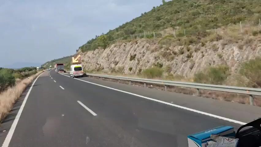 A highway exists with a steep, rocky, and vegetated hillside on the right. A lane of traffic moves away from the viewer. A white van with flashing yellow lights on top and reflective markings on the back is driving in the right lane. A white truck with a red cover over its trailer is in the left lane, passing the van. The sky is overcast with some sunlight breaking through. The camera appears to be inside another vehicle, with a partially visible box and other items in the foreground.