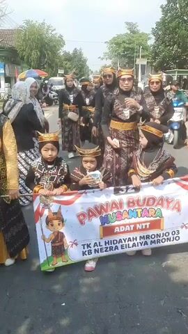 A group of people in traditional attire are participating in a parade. In the foreground, several children in elaborate headdresses and patterned clothing are holding a banner that reads "PAWAI BUDAYA NUSANTARA ADAT NTT TK AL HIDAYAH MRONJO 03 KB NEZRA EILAIYA MRONJO". Behind them, a larger group of adults, also in traditional clothing, are walking down a paved street. Some of the adults are wearing hijabs. The background shows a street with trees, buildings, and several vehicles, including motorcycles and cars. The overall atmosphere appears festive.