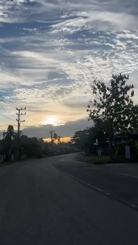 The video shows a road with some buildings and trees on the side. There are power lines running overhead. The sun is setting or rising in the distance, casting a warm glow across the sky. There are a few vehicles on the road, including what appear to be motorcycles. The overall impression is a rural or semi-rural scene at dawn or dusk.