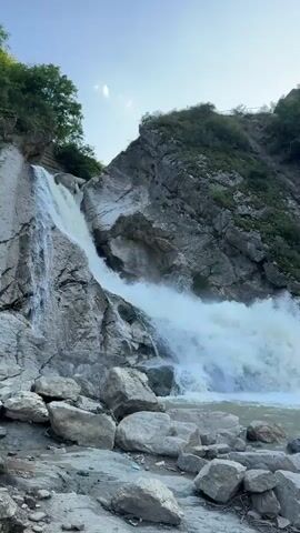 The video shows a powerful waterfall cascading down a rocky mountainside. The water is white and frothy as it plunges into a pool below. The rocks around the waterfall are rough and uneven, with some large boulders scattered in the foreground. Green vegetation covers the upper slopes of the mountain. The sky above is a clear blue with some wispy clouds. The overall impression is one of natural beauty and raw power.