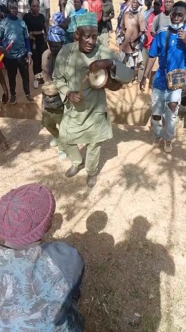 The video captures an outdoor gathering of numerous people, predominantly men, on dry, dusty ground under a clear, sunny sky, indicated by the shadows. In the foreground, a man dressed in a green outfit and a green and white patterned cap plays a small hand drum with a stick, contributing to the music. Another man in a light blue/green traditional outfit is also seen playing a similar drum in the mid-ground. A crowd of people stands around, observing the drummers and the event. Notable details include one person in the crowd wearing a red and white Santa-like outfit, and another in the foreground wearing a red knitted hat. The overall scene suggests a cultural or celebratory event, possibly in Kagoro, Nigeria, centered around live drumming. No visible text is present.