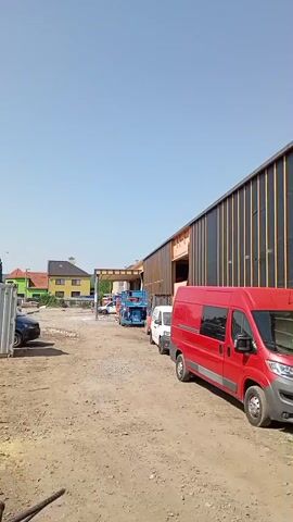 This media file shows a construction site with a partially built building. In the foreground, a red van is parked on a dirt road. Behind the van, a long, dark-colored building with vertical wooden beams is under construction. Scaffolding is erected along the side of the building, and construction materials are scattered on the ground. Further in the background, a yellow house and trees are visible, suggesting a semi-rural or suburban setting. The sky is clear and blue, indicating a sunny day, likely in the late morning or early afternoon. The overall scene depicts ongoing construction work on a clear, pleasant day. A license plate on the red van reads "5Z7 7073".