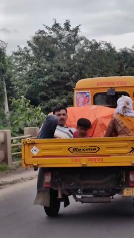 The video shows the back of a yellow autorickshaw, possibly a three-wheeled vehicle. There are three people visible in the back. A man with dark hair and a mustache is looking towards the camera. Next to him, a young child with dark hair is also looking at the camera. To the right, a woman with a white cloth covering her head and a yellow and orange sari is visible. The back of the autorickshaw is covered with an orange tarp. A sticker with images of people is on the top right of the tarp. The vehicle appears to be on a road with greenery and trees in the background. The words "MAXIMA" and "जय भगवान" are visible on the side of the autorickshaw.