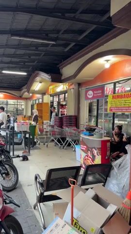 The video shows the exterior of a small supermarket. In the foreground, there are several parked motorcycles, and a few chairs with boxes and brooms piled behind them. The entrance to the supermarket is visible, with shopping carts lined up outside. Inside the store, several people are seen browsing or working. One person in a hijab is looking at products on a shelf. Another person stands near the checkout area. A yellow sign with red text is displayed near the entrance. The overall environment appears to be a somewhat cluttered and busy commercial area.