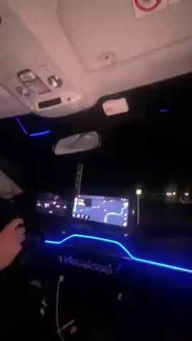 The video shows the interior of a car at night. The dashboard is lit with blue LED lights. There is a navigation system on the center console screen. A person is seen holding the steering wheel and driving the car. The environment is dark and only visible through the windshield are streetlights and passing cars.