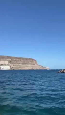 The video shows a view from a boat or watercraft moving across the ocean on a clear day with a bright blue sky. The camera moves, panning from left to right. A rocky promontory is visible on the left side of the frame. As the camera pans right, a man-made structure becomes visible, consisting of a breakwater made of large rocks and a building with a white exterior and a red-and-white striped tower element. Further to the right, a pier is visible, populated with several white buildings and a yellow attraction. People are on the pier. The water is a dark blue color.