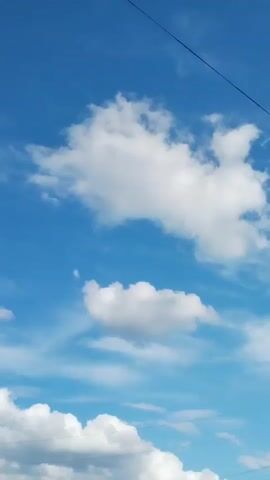 The video shows a clear blue sky with scattered white clouds. There are some power lines visible in the frame, as well as a utility pole. Trees can be seen at the bottom of the frame. The camera is pointed upwards, capturing the expanse of the sky. The footage appears to be taken outdoors during the daytime.