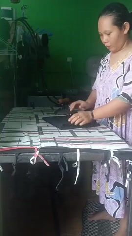 A woman is ironing clothes on a table. She is wearing a purple and white patterned dress and has her hair pulled back. The ironing board is covered with a patterned cloth. An iron is on the board, and steam is coming from it. The woman is holding the iron with her right hand and appears to be smoothing out a black piece of clothing with her left hand. The background is a green wall.