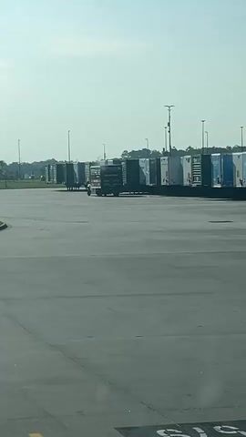 This is a video file shot from a vehicle's perspective, looking out at a wide, paved area, likely a truck yard or distribution center. The scene is dominated by a large number of parked semi-trailer trucks, lined up in rows under a clear, bright sky. The sun appears to be high in the sky, suggesting it's daytime, possibly mid-morning or afternoon. The weather seems fair, with no visible precipitation or heavy cloud cover.

In the mid-ground, a single vehicle, likely a small truck or utility vehicle, is moving amongst the parked trailers. It is difficult to discern the exact activity, but it could be repositioning trailers, performing checks, or engaged in logistical operations within the yard.

The trailers themselves are varied in color and branding, though many appear to be white or blue. Some have visible logos or text, including what looks like the Amazon logo on a few trailers. In the distance, there is a line of trees and buildings, suggesting the location is on the outskirts of a town or city. The foreground shows pavement with yellow and blue markings, indicating parking or driving lanes.

There is no discernible emotion conveyed in the video, as it focuses on the industrial setting and the vehicles within it. The location appears to be a functional hub for logistics and transportation, characteristic of areas where goods are stored and distributed.