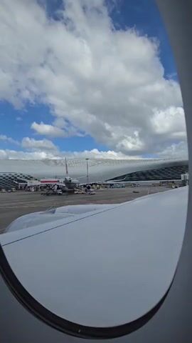 This video shows a view from an airplane window. The airplane is on the tarmac at an airport. The wing of the airplane is visible in the foreground, taking up the right side of the frame. In the middle ground, there are several other airplanes parked on the tarmac, as well as airport buildings and infrastructure. The sky is blue with some white clouds. The video is static, with no movement or action occurring.