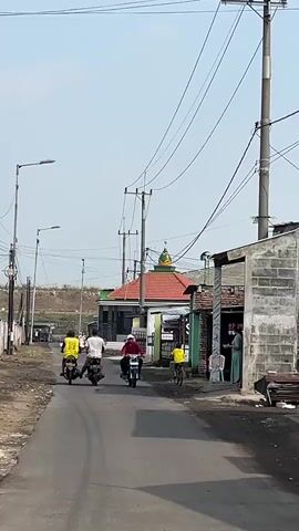 The video shows a rural street scene under a clear sky. A paved road runs through the center of the frame, with two motorcyclists and one cyclist riding away from the camera. On the right side of the road, a woman in a green dress stands outside a small shop. Further down the street, a mosque with a green dome and red roof is visible. Power lines crisscross the sky, supported by wooden poles. The environment appears to be a mixture of residential buildings and open land.