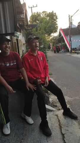 The video opens with a person in a red shirt and black pants sitting on a concrete ledge, giving a thumbs up to the camera. They are wearing a black baseball cap and smiling. To their left, another person in a red shirt and black pants is also visible, smiling. The background shows a street decorated with Indonesian flags, suggesting a national celebration. Several other people in red shirts and white pants are seen walking and standing along the street, with trees and buildings lining the road. The lighting suggests it is late afternoon or early evening. The camera pans to show more of the street scene, including more people gathered and a stage-like structure in the distance. The overall atmosphere appears festive and communal.