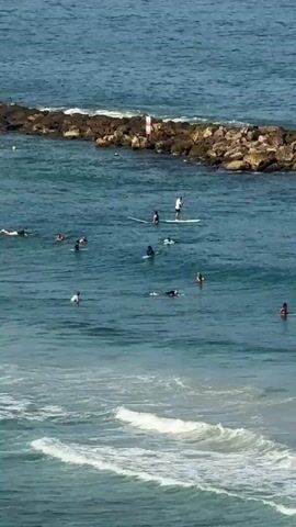 The video shows a body of water with several people engaged in water activities. In the foreground, waves are breaking and rolling towards the shore. In the midground, multiple individuals are on surfboards, either paddling or lying on them. One person is standing on a paddleboard. Further in the background, a stone breakwater extends into the water, with a red and white marker buoy visible on it. The water is a clear blue, and the sky appears bright, suggesting a sunny day. The overall scene depicts a recreational setting at the beach.