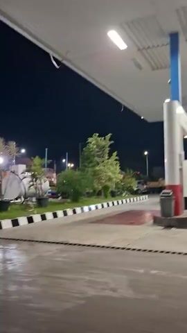 The video shows a night scene at a gas station. The camera is angled low and appears to be moving slightly. A black SUV is driving past the gas station sign, which displays the "PERTAMINA" logo and fuel prices. There are also trees and plants in potted containers around the gas station. The ground is wet, suggesting it might have rained recently. The lighting suggests it is nighttime, with artificial lights illuminating the area.