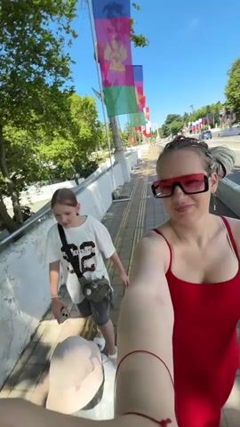 This is a selfie-style video captured on a bright, sunny day, likely in the afternoon given the strong shadows. The scene is an outdoor pedestrian walkway in Sochi, Russia.

The main subjects are three people. In the foreground, a woman wearing a red tank top and large, red-tinted sunglasses smiles at the camera. She has her hair in styled braids. Behind her and to the left, a young girl with blonde hair, wearing a white t-shirt with a graphic design and grey shorts, walks along the path. She is looking at something off-camera and seems engaged in a conversation. Partially visible in the bottom of the frame is a very young child wearing a light-colored hat and a grey outfit.

The walkway is paved with yellow tactile paving tiles, indicating a public space, possibly near a road or waterfront. A low, white, textured wall runs along the left side of the path. Behind this wall, lush green trees with visible branches create a canopy overhead. Further back, a road with cars is visible, suggesting an urban or semi-urban environment.

A notable detail is a series of flags mounted on poles lining the walkway. These flags, in various colors including green, purple, and red, and featuring graphic designs, are characteristic of official or decorative displays often seen in public areas in Russia. The overall atmosphere appears cheerful and relaxed, with the subjects seemingly enjoying an outing. The text "SAN FRANCISCO" is visible on the young girl's t-shirt.