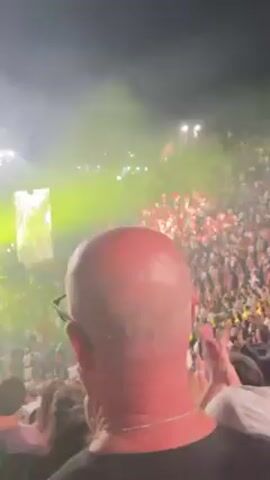 The video shows a concert at night. A stage is visible in the distance with a large screen displaying a performer. Green and yellow spotlights shine across the crowd. A large crowd is visible in front of the stage, with many people raising their hands and cheering. A person with a shaved head is standing in the foreground, partially obstructing the view of the stage. The environment appears to be a large outdoor venue.