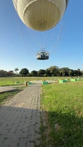 Here's a description of the video:

The video shows a large, tethered hot air balloon hovering above a park-like area. The balloon is primarily white with black markings and the word "MORE" in large, green letters and some Hebrew script. A metal basket, likely for passengers, is suspended beneath the balloon. The sky is clear and blue. The ground below has a brick pathway leading towards the balloon. Grass, trees, and some small green and yellow structures are also visible. The balloon appears to be stationary.