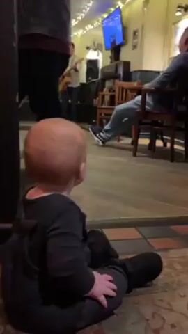 A baby sits on the floor of a bar, facing away from the camera, watching a man play guitar. The bar has a television screen, a tiled floor and wooden chairs. Another person sits in a chair in the background. One person stands between the baby and the performer.