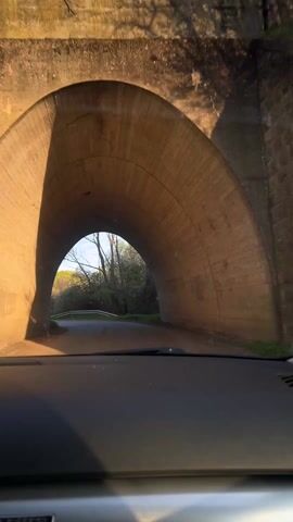 A car drives out of a concrete tunnel into a wooded area. The trees are mostly bare.  The car drives along a narrow, winding road bordered by a guard rail. The sky is blue with some clouds.