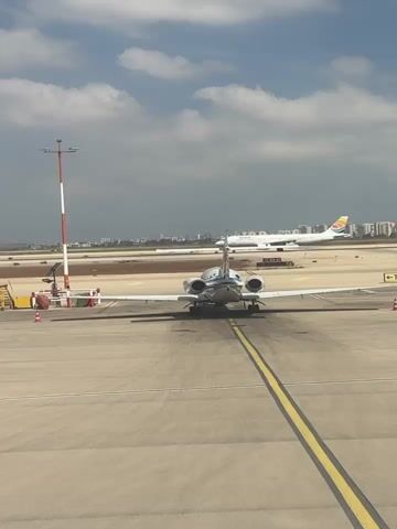 The video shows an airport scene with two airplanes. A smaller private jet is in the foreground, facing away from the camera on a tarmac. In the background, a larger commercial airplane is on a runway, also facing away from the camera. There is a white truck with a logo on it between the two airplanes. The sky is partly cloudy. The overall environment is that of an active airport.