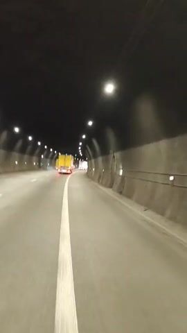 This media file shows a view from inside a tunnel. The tunnel has concrete walls and a dark ceiling with rows of lights. The road surface is asphalt and marked with a white line dividing lanes. A yellow truck is visible in the distance, driving away from the camera. A black car is seen to the left of the frame, also driving in the same direction. The overall scene suggests a vehicle traveling through an underground passage, likely during daytime as indicated by the lighting in the tunnel. There are no visible people or explicit location cues other than the provided context. The activity is transportation.