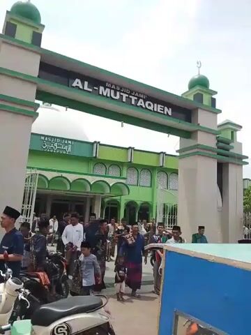 The video shows a green mosque named "Al-Muttaqien" with people in traditional Muslim attire gathering outside.  Some people are walking, others are getting on or off motorbikes.  There are many motorbikes parked in front of the mosque, and a blue vehicle, possibly a food cart, is also present. The sky is cloudy.