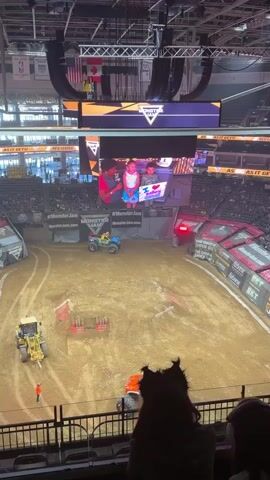 The video shows an aerial view of an indoor arena set up for a monster truck event. The floor is covered in dirt with obstacles and vehicles. In the center of the arena, there are several vehicles, including a yellow bulldozer, an ATV with a rider, and a monster truck. Spectators are seated in the stands around the arena. A large video screen suspended from the ceiling displays images of children. The overall atmosphere appears to be a live sporting event.