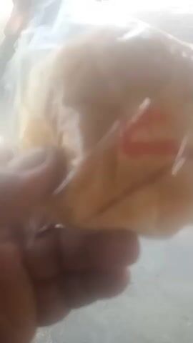 A person's hand, with visible fingernails, holds a round, light-brown food item, possibly a bun or pastry, wrapped in clear plastic. The item appears to have a red marking on its surface, possibly a stamp or decoration. The background is blurred but suggests an outdoor environment with a paved surface in the foreground and a red motorcycle with a license plate visible in the mid-ground. Sunlight seems to be casting a bright light on the scene, creating some overexposure.