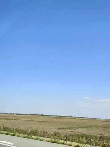 A wide shot captures a vast, flat, dry field under a clear blue sky. The field is sparsely vegetated, with some evidence of tilled rows. In the distance, a large agricultural irrigation system, likely a center pivot irrigator, stretches across the landscape, its metal arms and wheels visible. The foreground features a road or path on the left and a fence line with wooden posts and wire. The overall impression is one of arid agricultural land on a bright, sunny day.