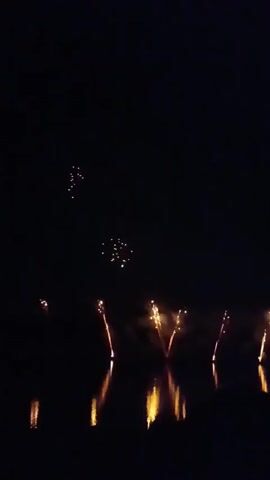 The video displays a nighttime scene with fireworks exploding in the sky. The fireworks are reflected in the water below, creating a shimmering effect. The colors of the fireworks are predominantly yellow and orange. The overall atmosphere is festive and celebratory.