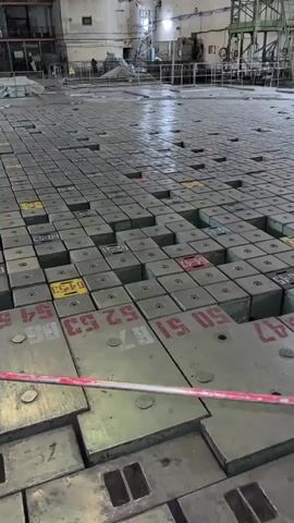 The video shows a wide shot of a vast indoor space, possibly a power plant or industrial facility, dominated by a floor covered in a grid of square blocks. The blocks are tightly packed and appear to be made of metal, with some bearing numbers and markings in red and yellow. In the background, there is a large, complex structure with multiple levels, scaffolding, and industrial equipment. The overall atmosphere is industrial and somewhat desolate, with the lighting suggesting an artificial, utilitarian environment. No people are clearly visible in the frame.