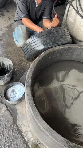 A person kneels on the ground next to a large, round tub filled with murky water. The person is holding a black tire with metal rims and appears to be inflating it with a tool connected to a hose. The tire is positioned on the ground next to the water tub. To the left of the tub, there are two smaller containers. One is a black bucket, and the other is a shallow metal dish containing a white substance. The ground appears to be wet and dirty, suggesting an outdoor or workshop environment. The person is wearing a black shirt and blue jeans.