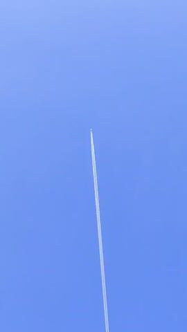 This is a close-up shot of a clear blue sky with a single airplane leaving a contrail as it ascends at a steep angle. The contrail is long and thin, indicating the plane is at a high altitude. The airplane itself is a tiny white speck at the very top of the contrail. The camera is angled upwards, focusing on the plane and its trail against the vast expanse of the sky.