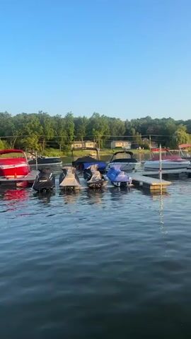 A tranquil body of water is seen under a clear blue sky, reflecting the serene surroundings. In the foreground, the gentle ripples of the water create a sense of calm. A dock extends into the water, lined with various watercraft. Several jet skis, covered for protection, are parked along the dock. Behind them, larger boats, some covered and some open, are also docked. The background features a lush green treeline, dotted with houses and power lines, suggesting a residential area near the water. The scene is bathed in the soft light of either early morning or late afternoon, casting a warm glow over the landscape. No people or animals are visible in the frame.